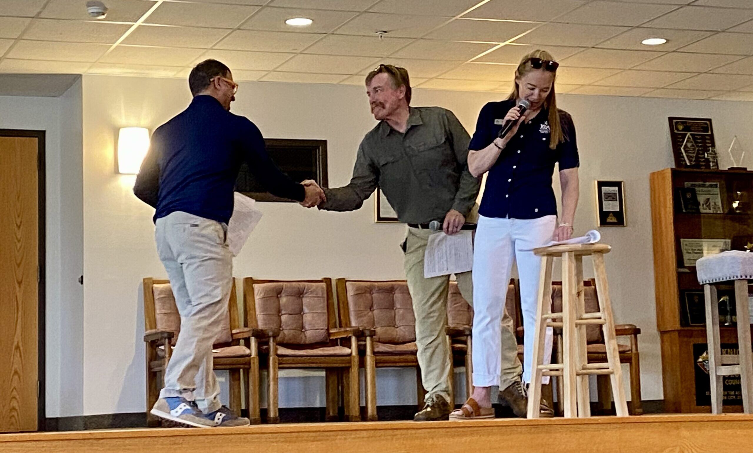 Emily Fisher, on the microphone, nine-time Olympic announcer; including Winter, Summer, Para, and Youth, and co-event emcee Carl Roepke to her left is seen shaking the hand in congralutations to their co-emcee Ryan 