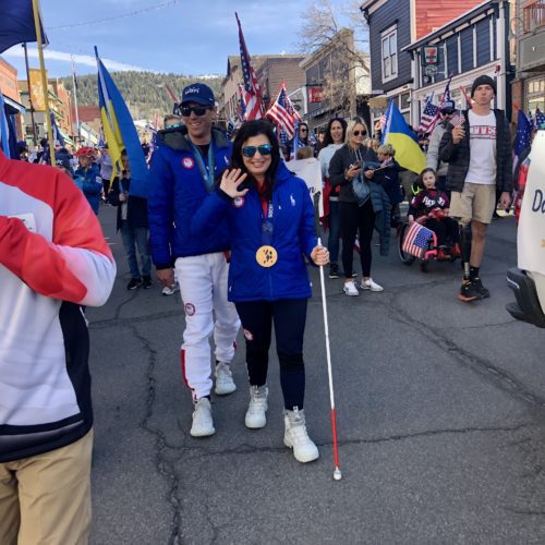 Visual impaired, alpine skier, Danelle and Rob Umstead (Guide and husband), flag bearer, 4-time Paralympian in blue. Nine-time Olympics announcer Carl Roepke, in white and red, announcing the Park City Nation, Youth Sports Alliance (YSA) Parade down Main St.