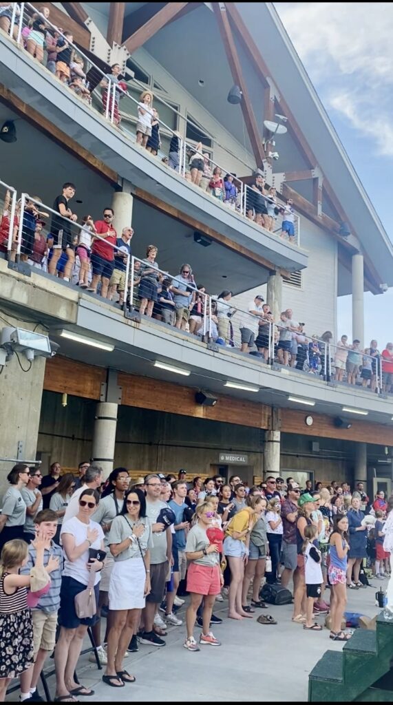 Rulon Gardner, bottom floor pool deck at Utah Olympic Park, standing in the back, wearing a dark blue short sleeve shirt, and holding his hand over his heart as the crown sang the National Anthem at the celebration for the announcement of Ytah's winning bid to host the 2034 Olympics.