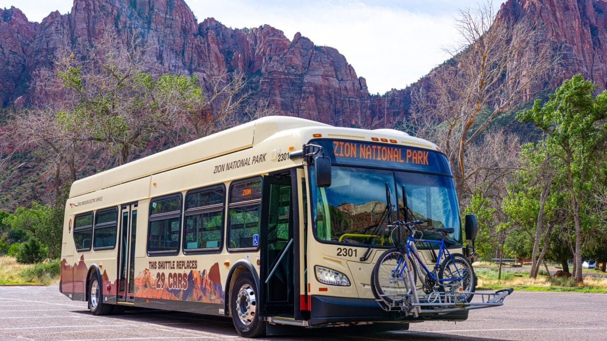 Electric shuttle bus with bikes on a rack mounted in front stands in Zion National Park with red rock towering behind. Text on bus reads, "This shuttle replaces 29 cars." A new electric shuttle bus in front of The Watchman in Zion National Park.