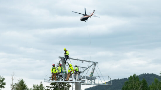 A blackhawk helicopter flies in the background and workers ready the sheave train of a newly installed tower on the Sunrise Gondola Project at the Canyons, Park City Mountain Resort.