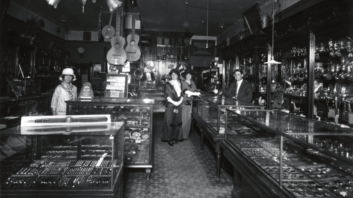 2005-27-18: Interior of Hodgson Jewelry store, ca. 1904-1914. Charles Hodgson stands behind a case, a woman in a white hat stands off to the left, and Kate and Mary Byrnes stand in the center near Hodgson.