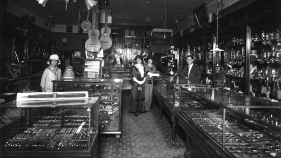 2005-27-18: Interior of Hodgson Jewelry store, ca. 1904-1914. Charles Hodgson stands behind a case, a woman in a white hat stands off to the left, and Kate and Mary Byrnes stand in the center near Hodgson.