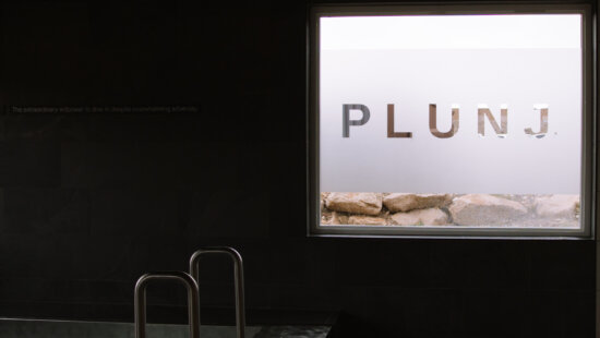 A view from inside PLUNJ Park City’s bathhouse, where the cold plunge sits beneath a frosted window bearing the studio’s name. The minimalist space reflects the Nordic-inspired simplicity of the hot–cold–rest ritual central to the PLUNJ experience.