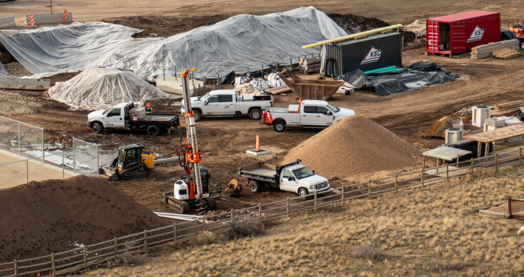 Elevated view of active construction and drilling at the Treasure Mountain School demolition site, with what appears to be a newly installed storm drain marked by an orange cone near the center of the image.