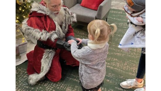 Santa kneels to greet a young visitor during a past holiday market at the DoubleTree – Yarrow Hotel in Park City, where families can enjoy free photos with Santa while shopping from local artists and small businesses.