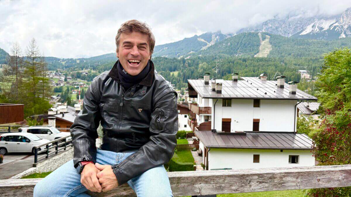 Kristian Ghedina sits outside Cortina's Hotel Trieste on September 13, 2025. In the background the Olimpia delle Tofane alpine skiing venue can be seen.