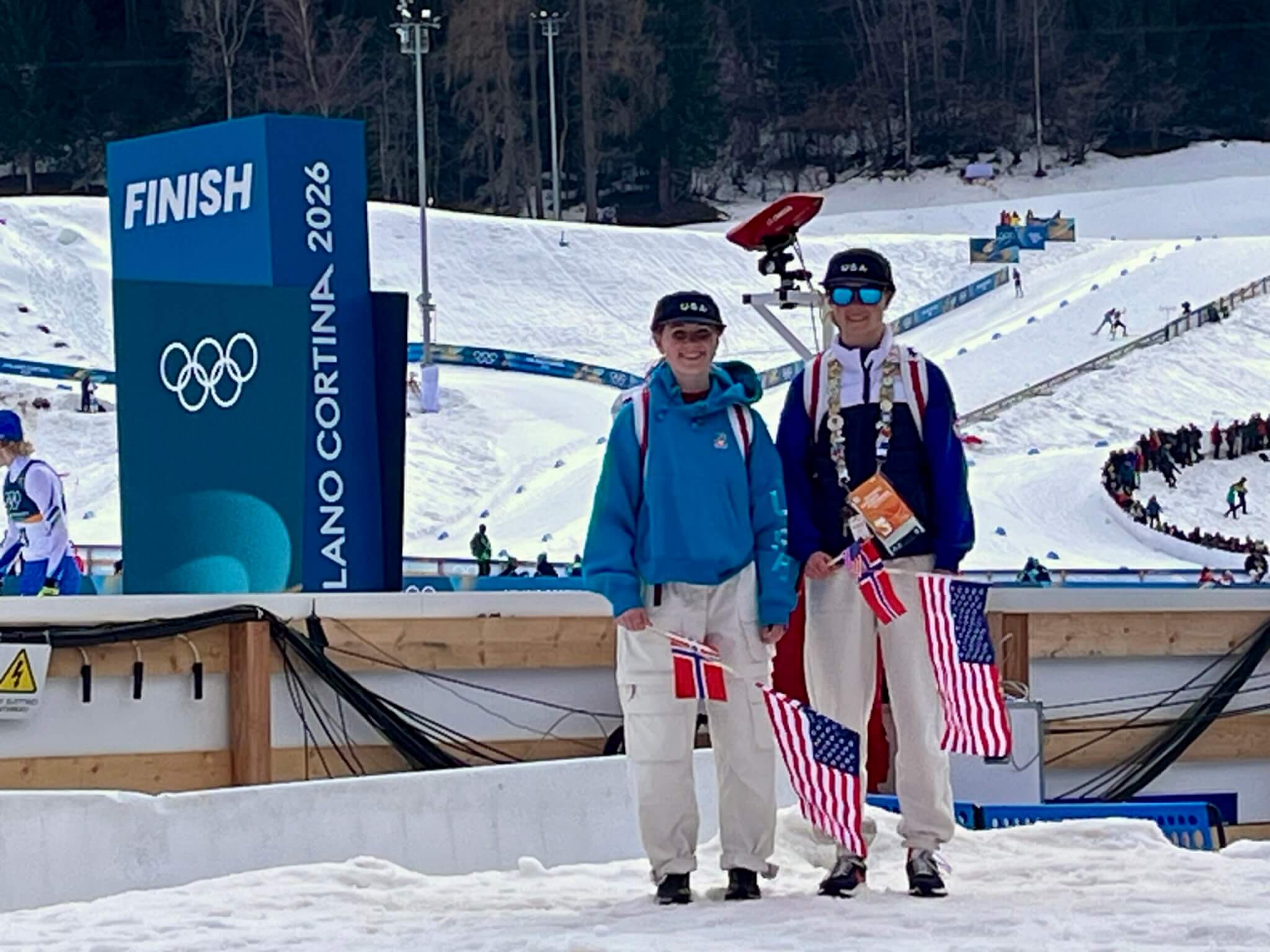 Josie Johnson and Annika Belshaw, Olympic USA women's Ski Jumpers In Milan Cortina cheering on their teammates, waving flags at the Finish Line.