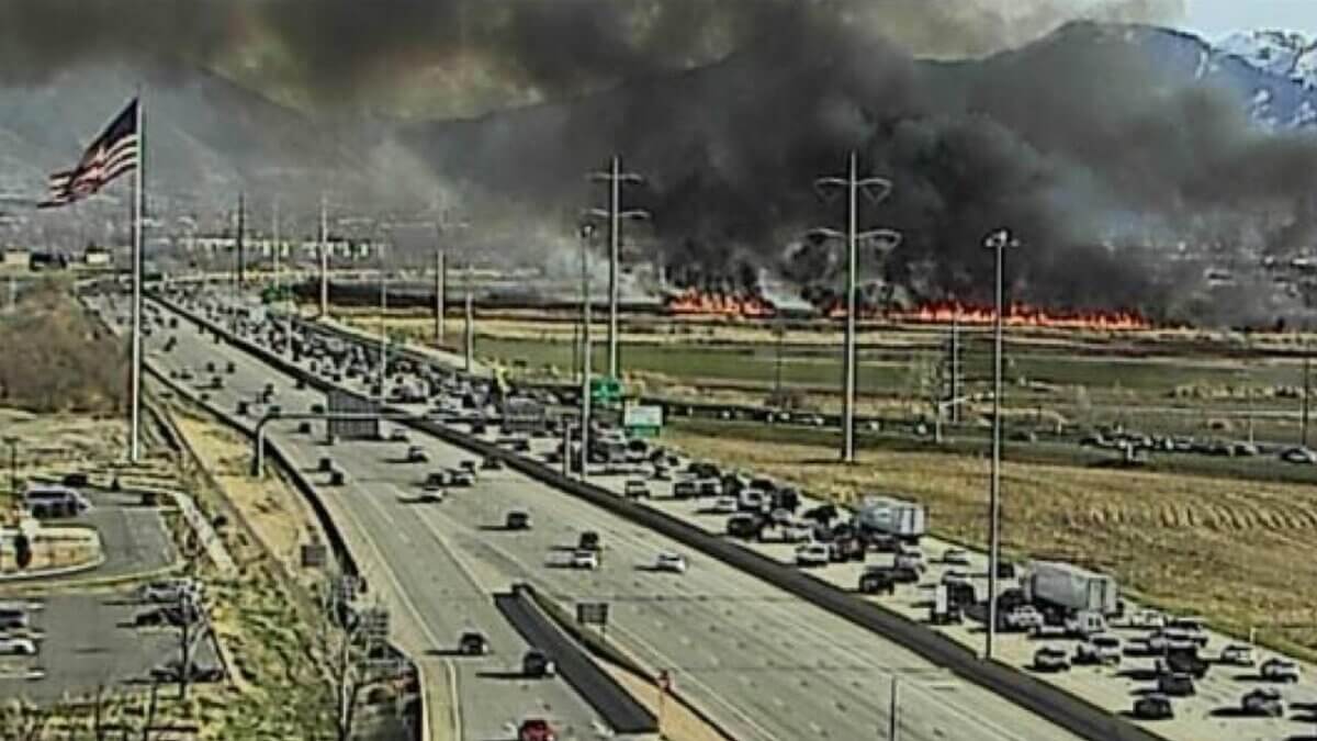 A UDOT traffic camera captures the Milrace Fire burning along the I-15 corridor in Utah County on Thursday, with heavy smoke rising above the Wasatch Front as traffic slows on the freeway below.