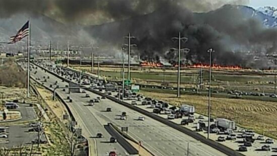 A UDOT traffic camera captures the Milrace Fire burning along the I-15 corridor in Utah County on Thursday, with heavy smoke rising above the Wasatch Front as traffic slows on the freeway below.