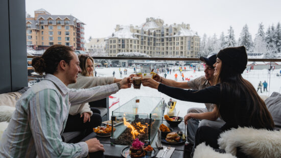 Guests share drinks and food around a patio fire table at the base of Whistler Blackcomb in February 2023.