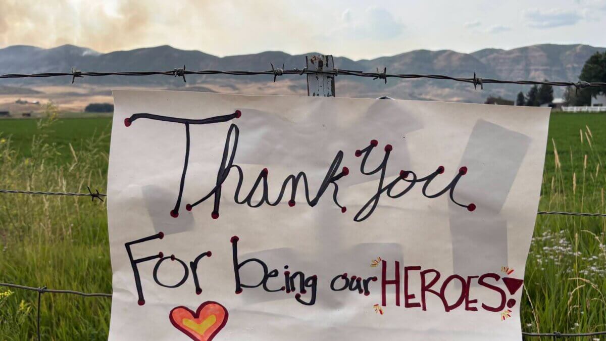 A handwritten sign reading "Thank you for being our heroes" hangs on a barbed wire fence as smoke from the Rocky Canyon Fire rises in the distance.
